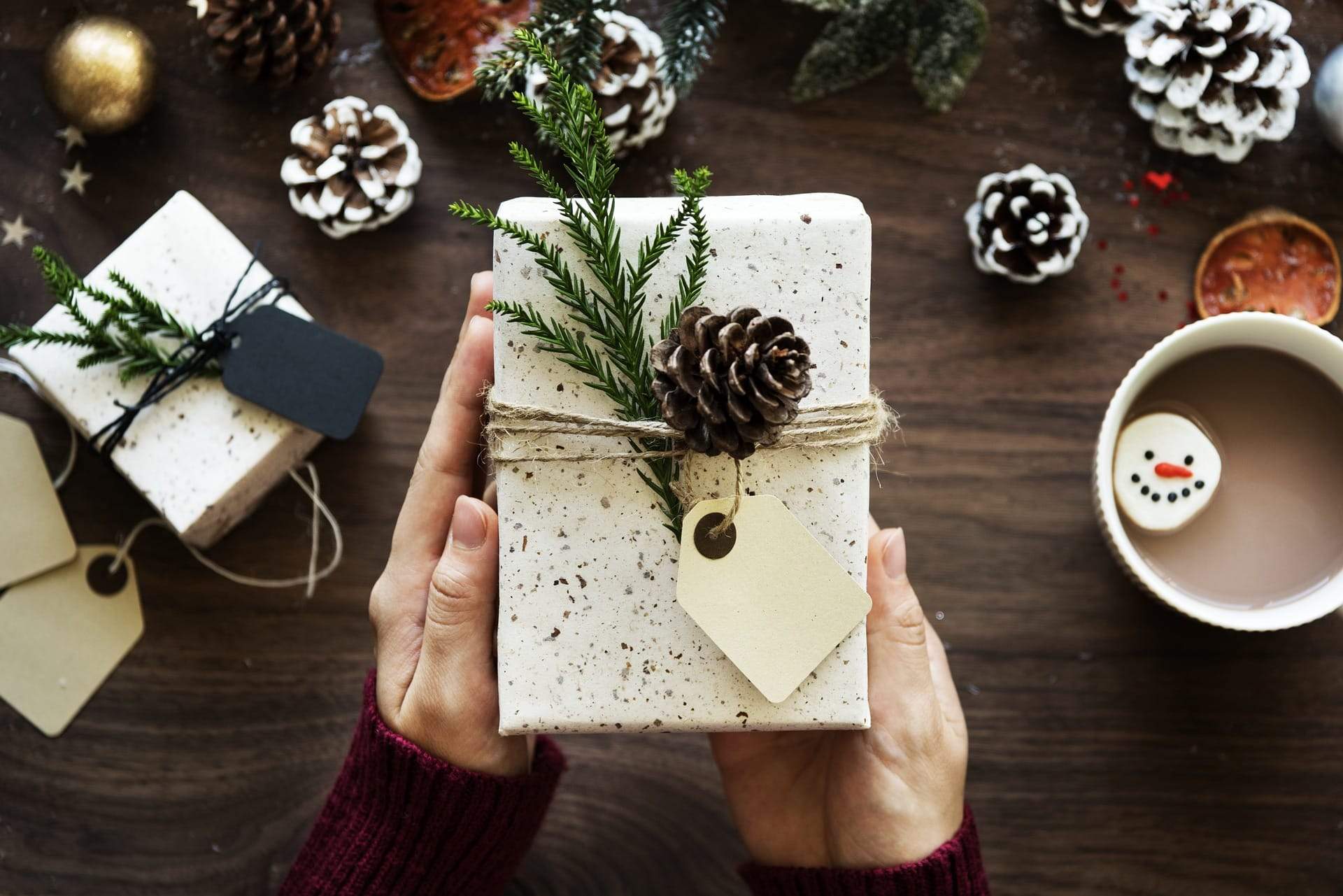 Hands holding a DIY gift descorated with a fern and pine cone; more pine cones and gifts in the background